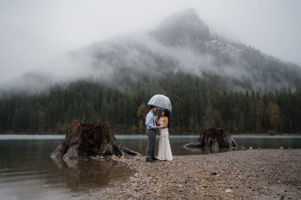 A couple who decided to elope in Washington State standing on the shore of Rattlesnake Lake, holding a clear umbrella.