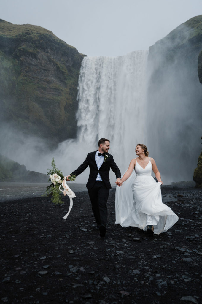 A couple who decided to elope in Iceland is running at the base of Skogafoss.