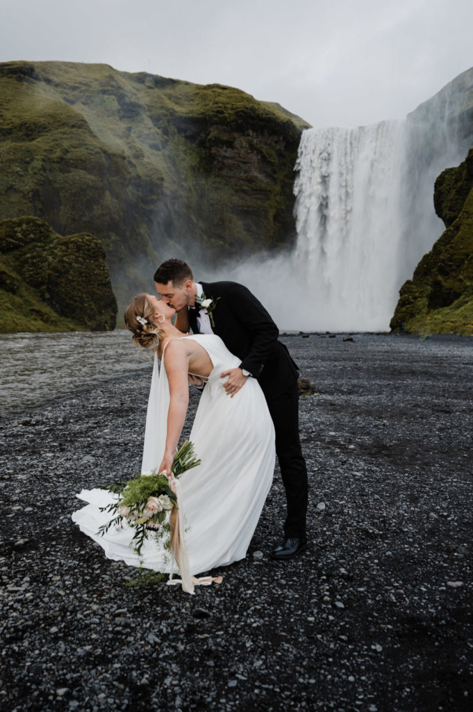 A couple who decided to elope in Iceland is standing in front of Skogafoss, kissing.