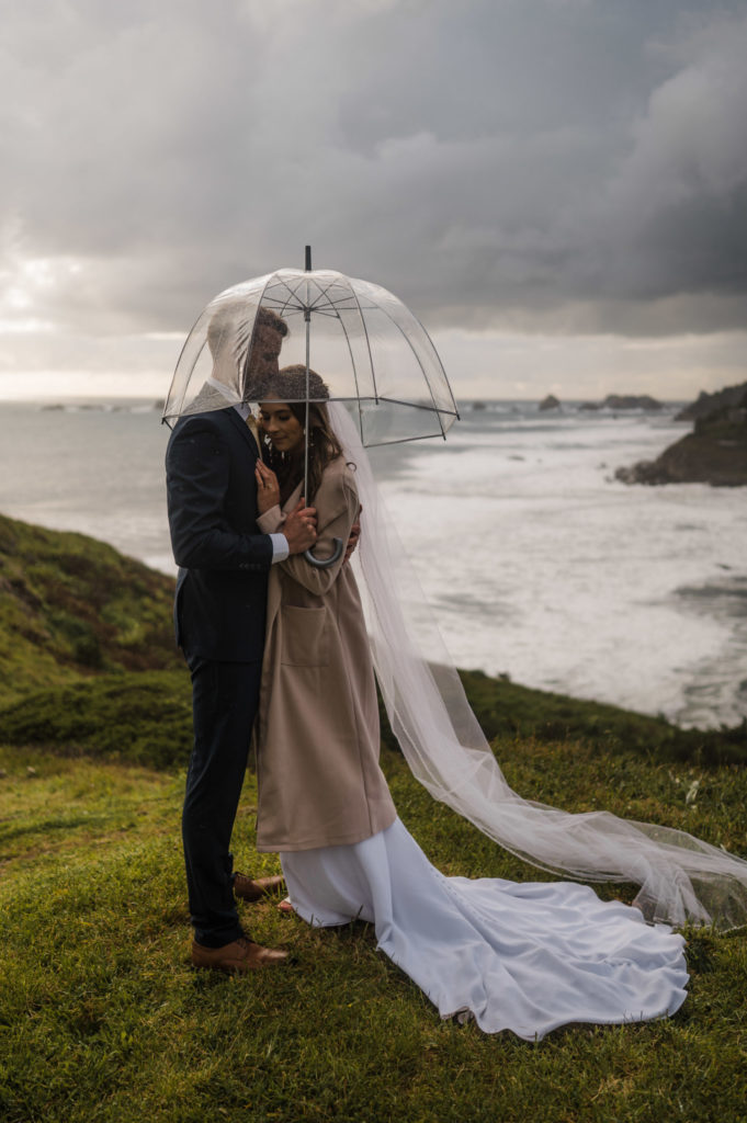 A couple standing under a clear umbrella next to the ocean, at one of the best places to elope in Oregon!