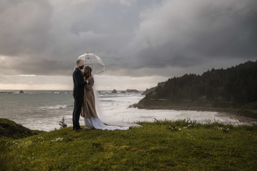 A couple standing under a clear umbrella next to the ocean, at one of the best places to elope in Oregon!