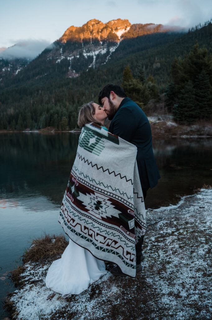 Couple wrapped up in a cozy blanket, kissing in front of Gold Creek Pond on their elopement day