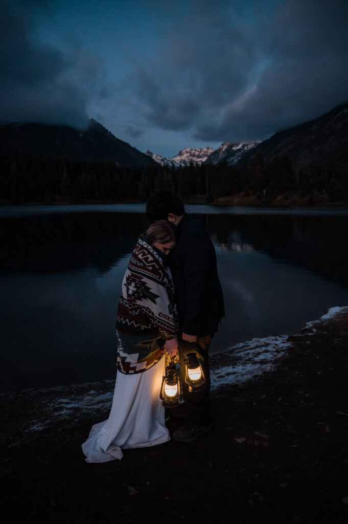 Couple cozied up under a blanket while holding lanterns at Gold Creek Pond in Washington on their elopement day