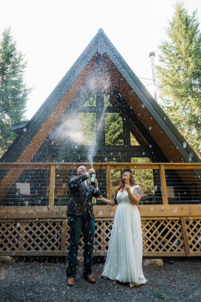 Couple popping champagne in front of their A-Frame cabin after eloping in Mount Rainier National Park 