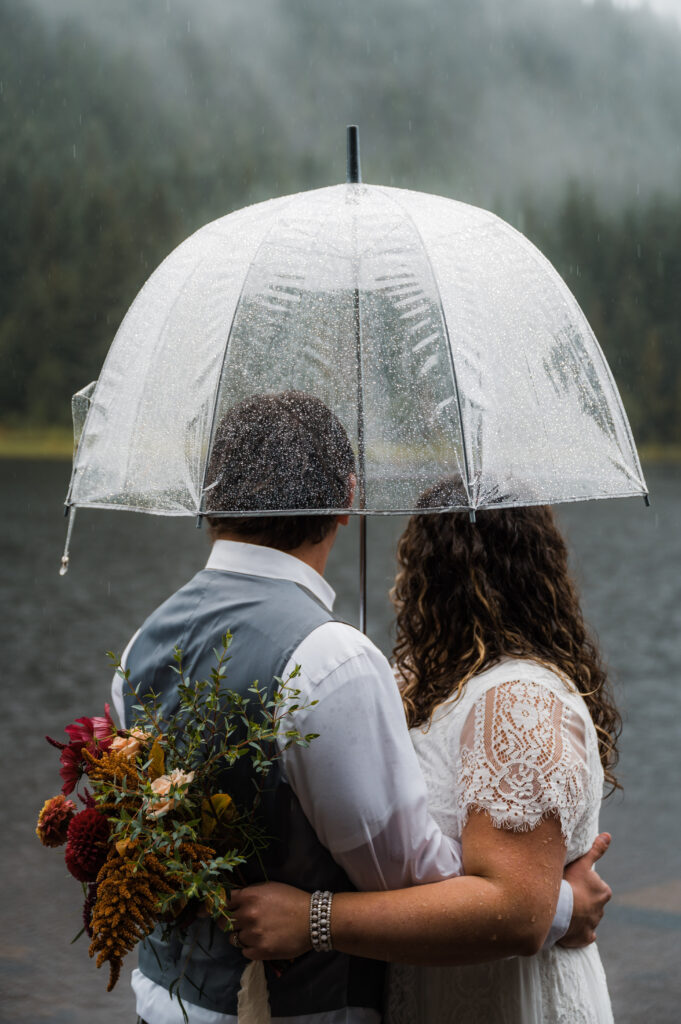 bride and groom standing under a clear umbrella in the rain during their elopement day at Trillium Lake in Oregon