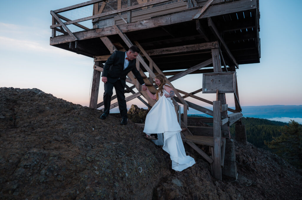 Groom holding hands with the bride and helping her up at a fire lookout in Washington
