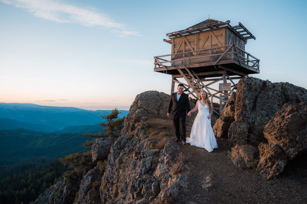 Eloping couple standing on the top of a cliff with a fire lookout in the background in Washington State