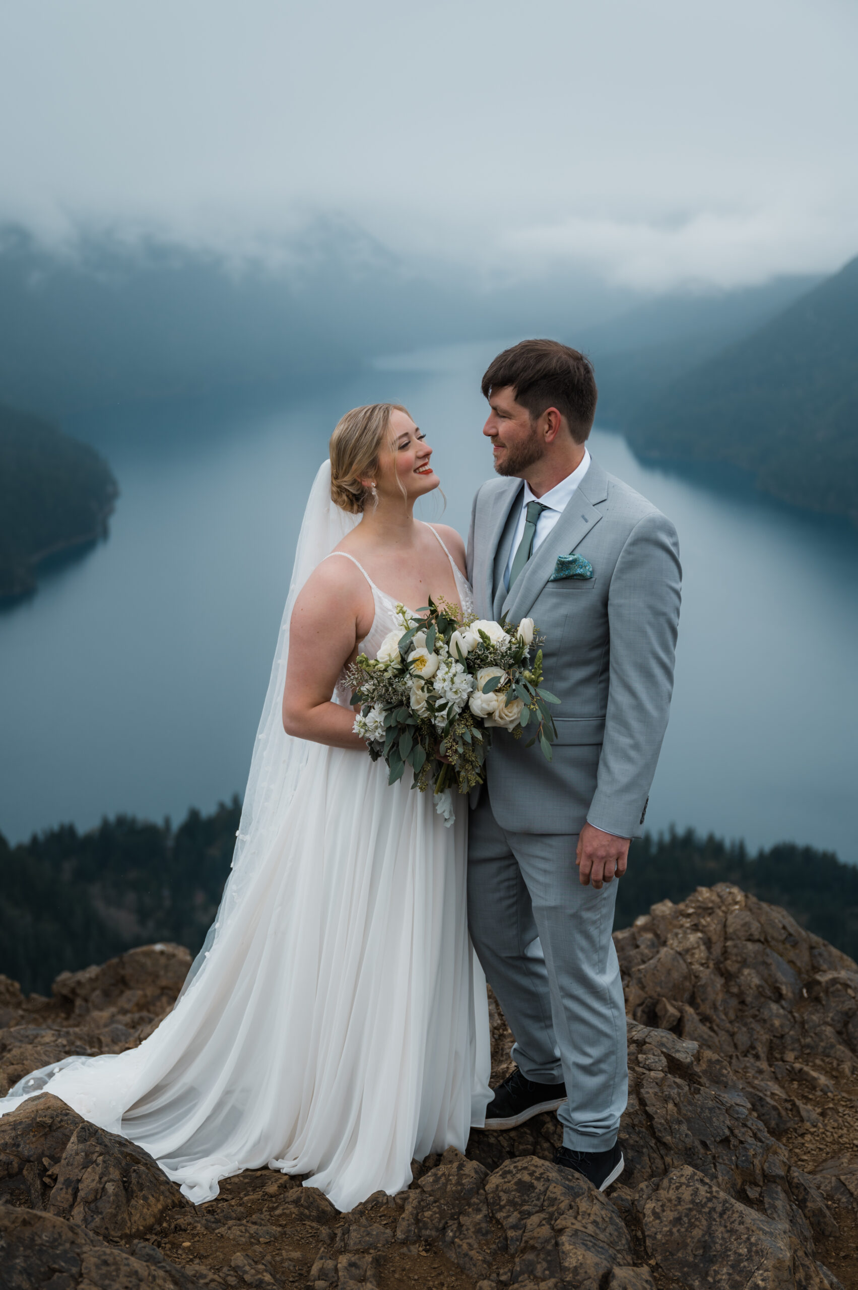 bride and groom looking at each other on top of Mount Storm King with views of the mountains and Lake Crescent in the background. Olympic National Park hiking elopement