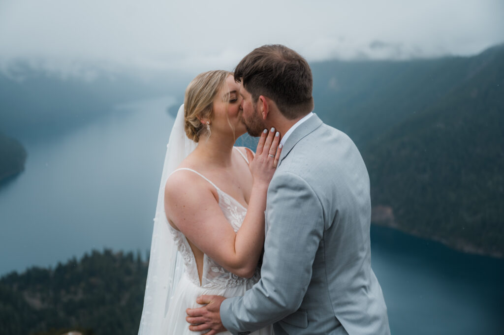 bride and groom sharing their first kiss during their hiking elopement on a foggy day with views of the mountains and Lake Crescent in the background