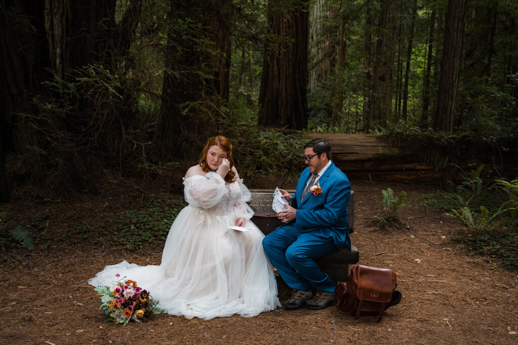 Bride and groom sit on a wood bench in Jedediah Smith Redwoods and read letters from family