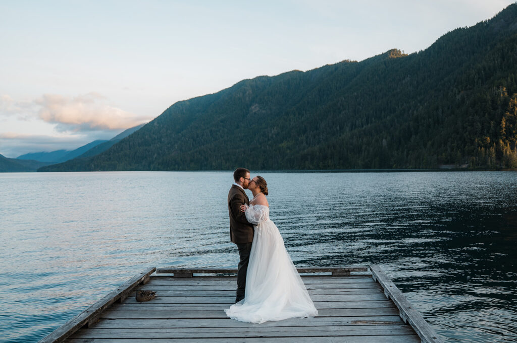 Bride and groom embracing on a wooden dock at Lake Crescent at sunrise, surrounded by forested mountains