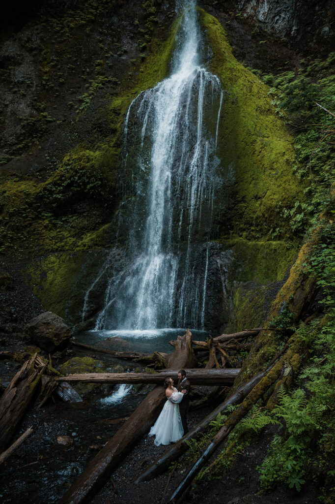 Bride and groom standing near Marymere Falls with cascading water in the background, smiling at each other.