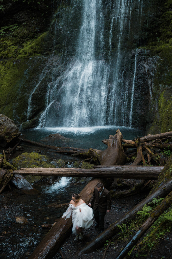 Bride and groom standing near Marymere Falls with cascading water in the background, smiling at each other.