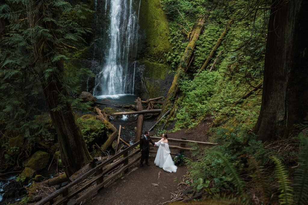 Bride and groom standing near Marymere Falls with cascading water in the background, smiling at each other.