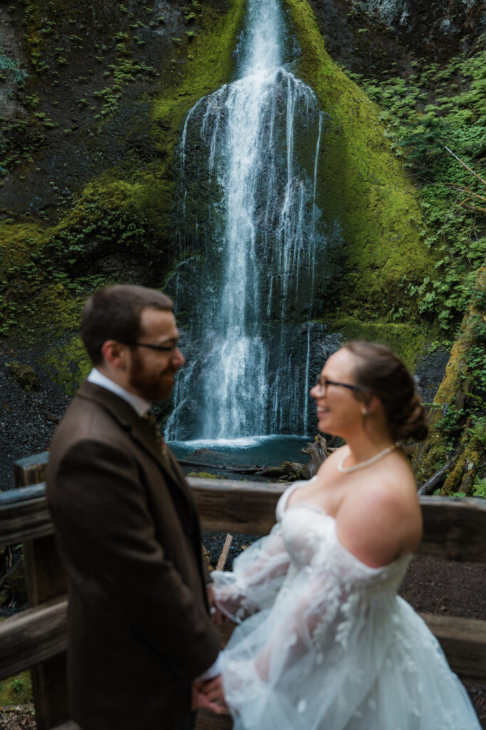 Bride and groom standing near Marymere Falls with cascading water in the background, smiling at each other.