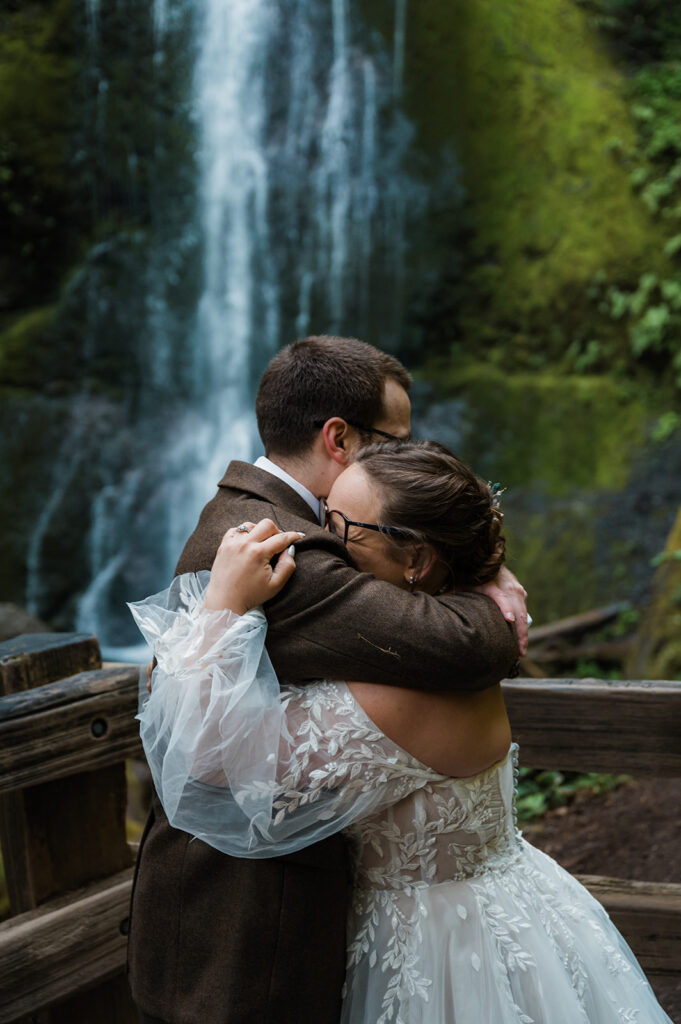 Bride and groom embracing near Marymere Falls with cascading water in the background