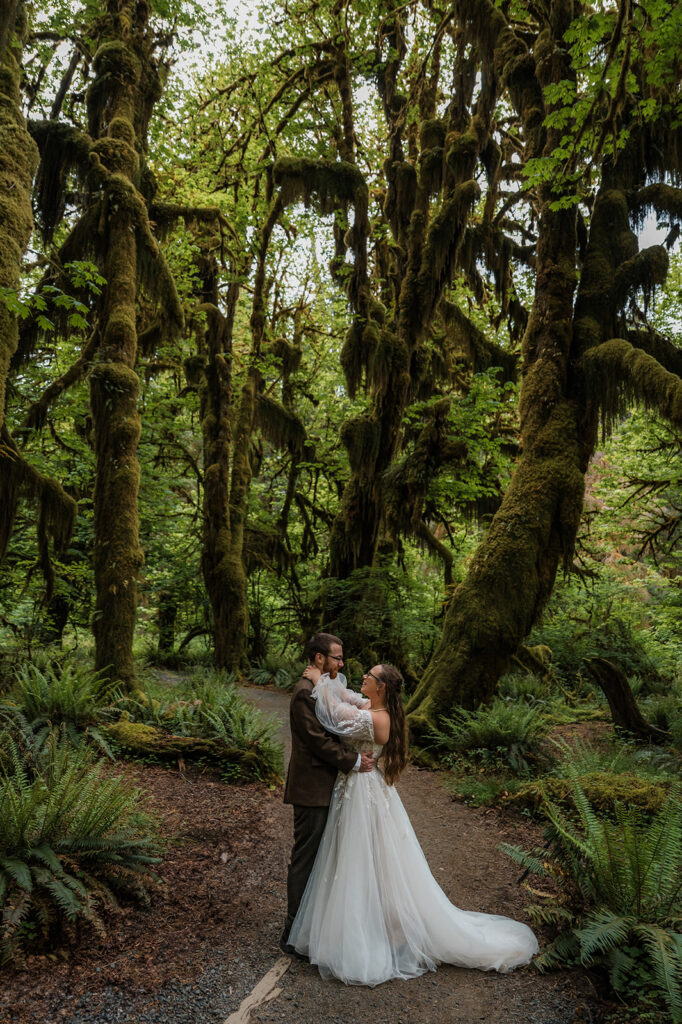 Bride and groom embracing in the Hoh Rainforest, surrounded by ancient trees draped in green moss
