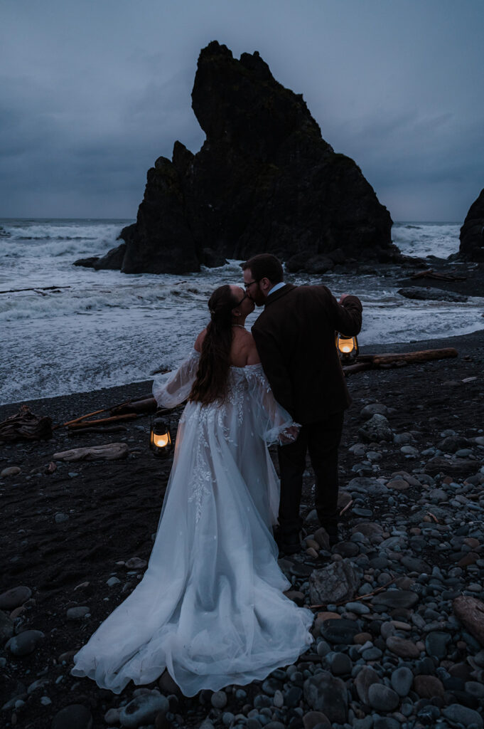 Bride and groom standing on Ruby Beach at dusk, holding lanterns, with the ocean and sea stacks in the background