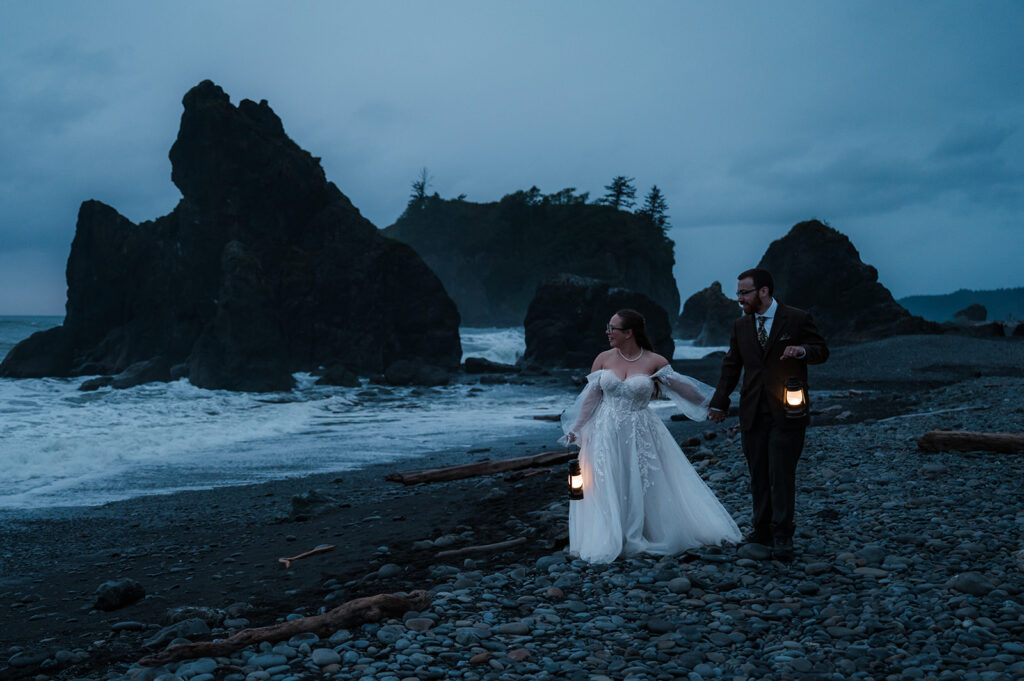 Bride and groom standing on Ruby Beach at dusk, holding lanterns, with the ocean and sea stacks in the background