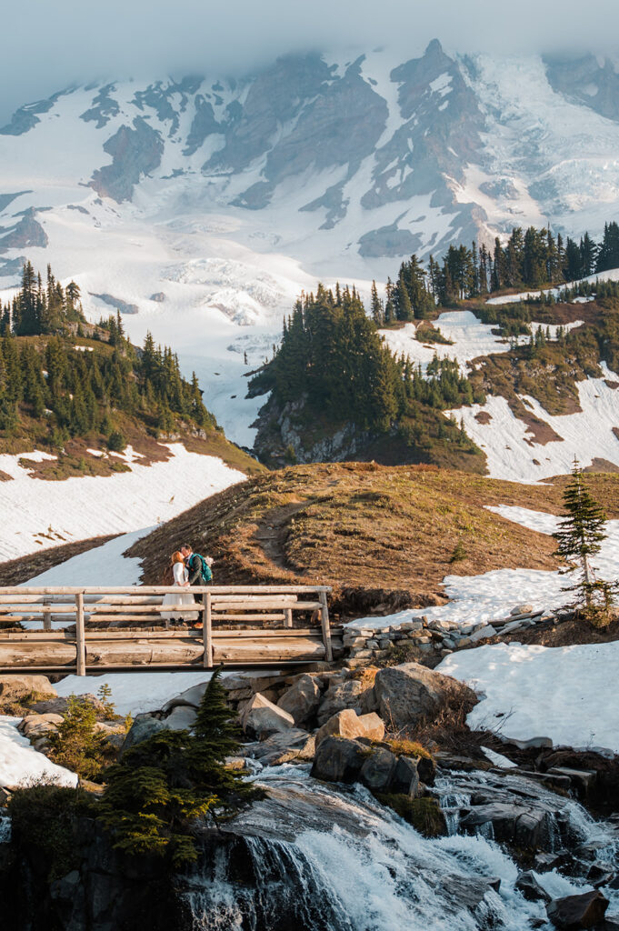 couple in their wedding attire standing on the bridge at Myrtle Falls with views of Mt. Rainier in the background on their elopement day